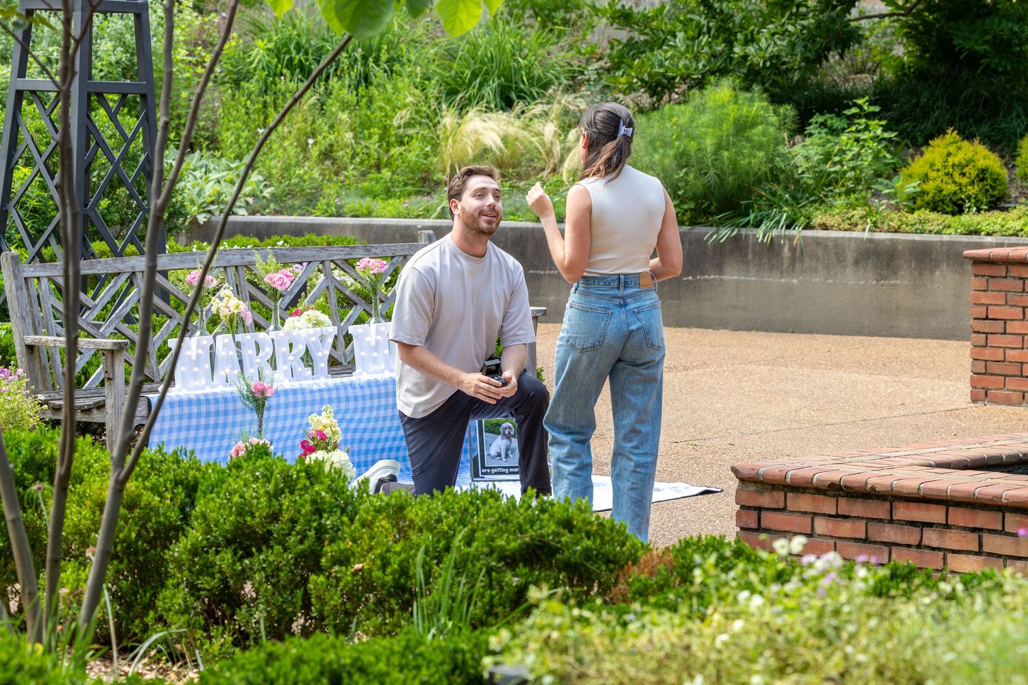 Man on one knee proposing to woman in a garden setting with floral decorations and lush greenery.
