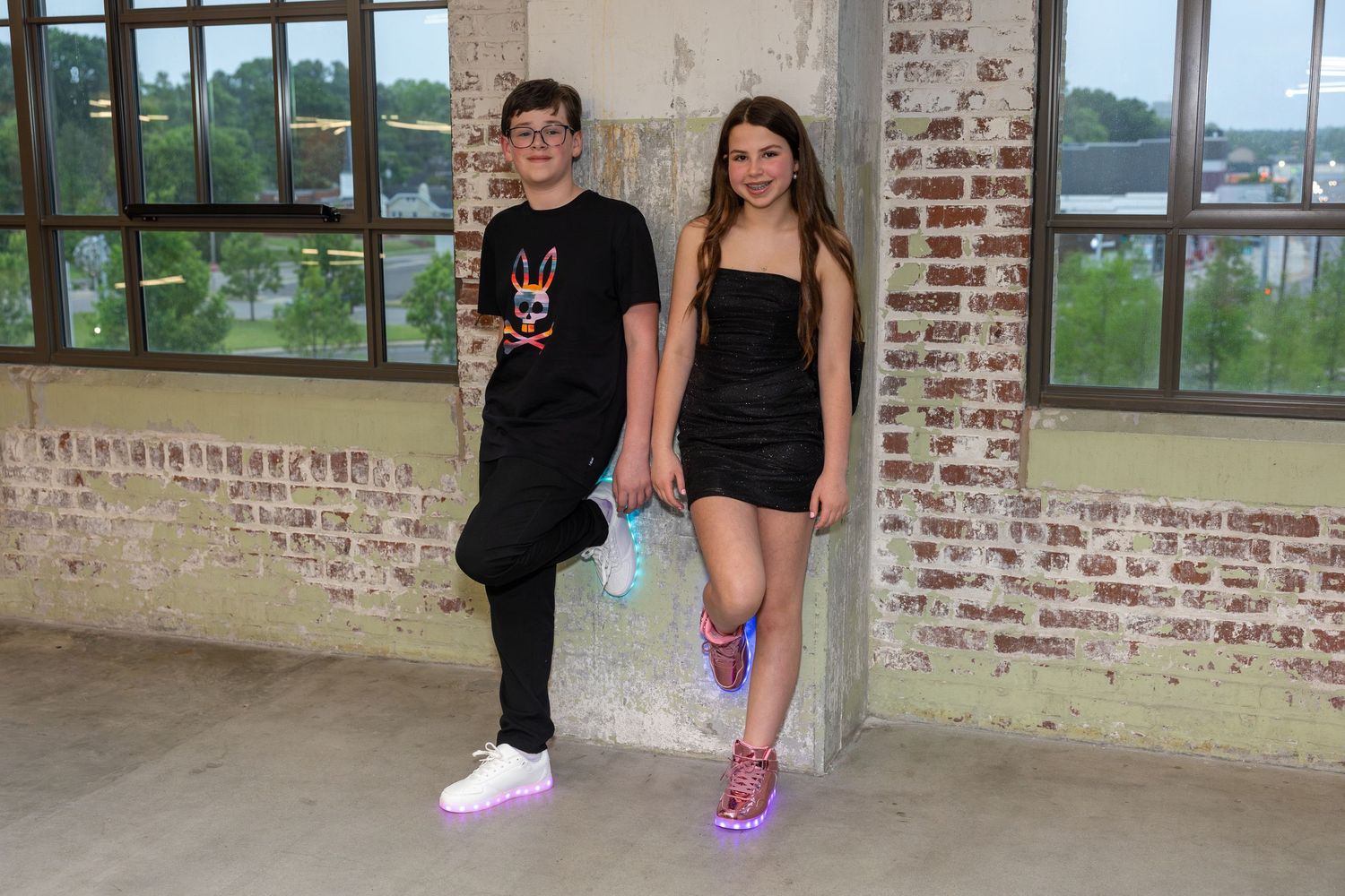 Two teens pose holding hands in a rustic brick building, wearing light-up sneakers and stylish outfits.