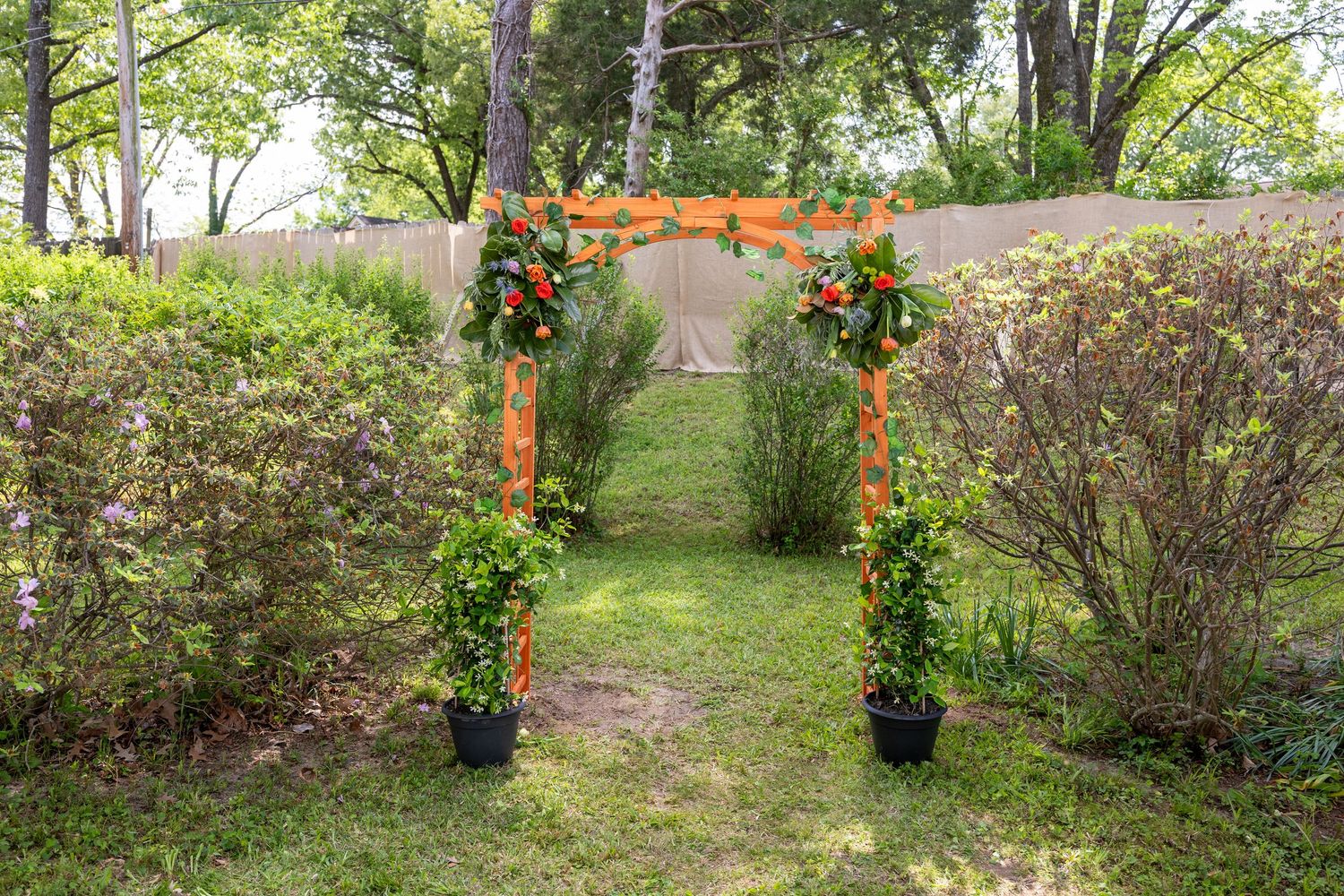 Orange floral wedding arch with potted plants on green lawn, garden ceremony backdrop with wooden fence.