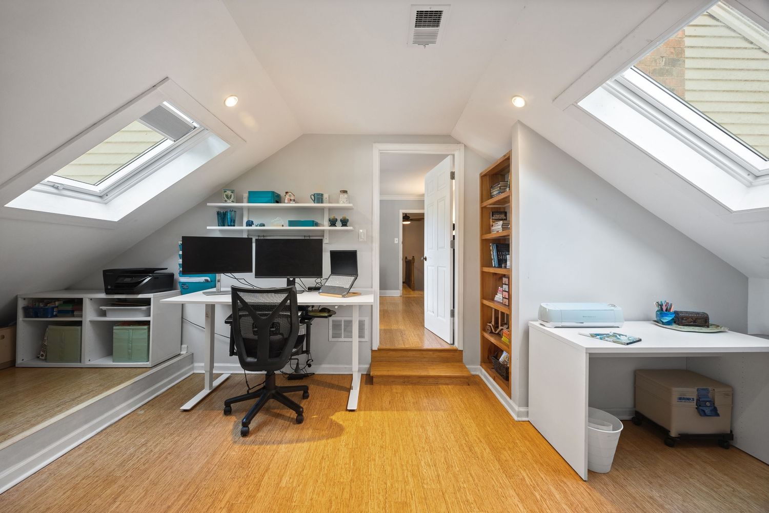 Modern home office in attic with skylights, bamboo flooring, built-in shelving, and white desk setup.