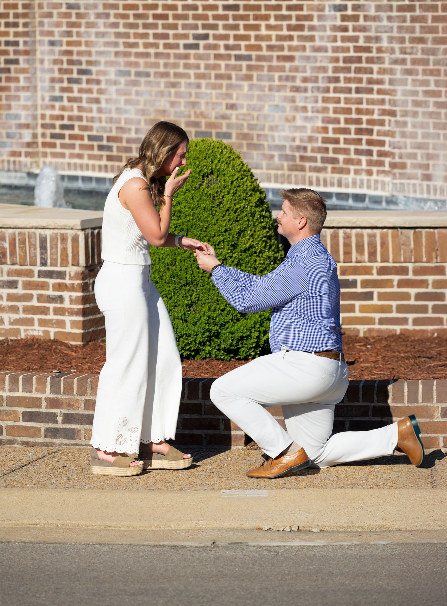 Man kneeling on one knee proposing to woman in white jumpsuit outdoors near brick wall and green shrub.