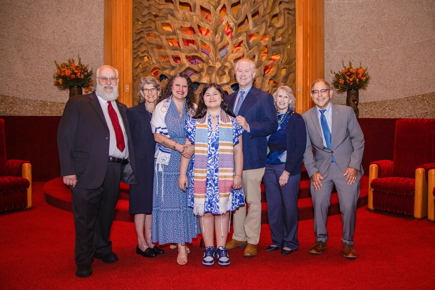 A group of people standing together in formal attire in front of an ornate wall with red carpeting in a grand room.