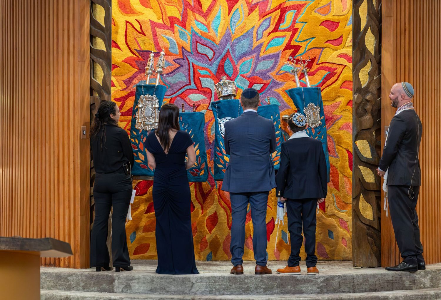 People standing before a vibrant colorful mural with teal Torah scrolls in a modern synagogue setting.