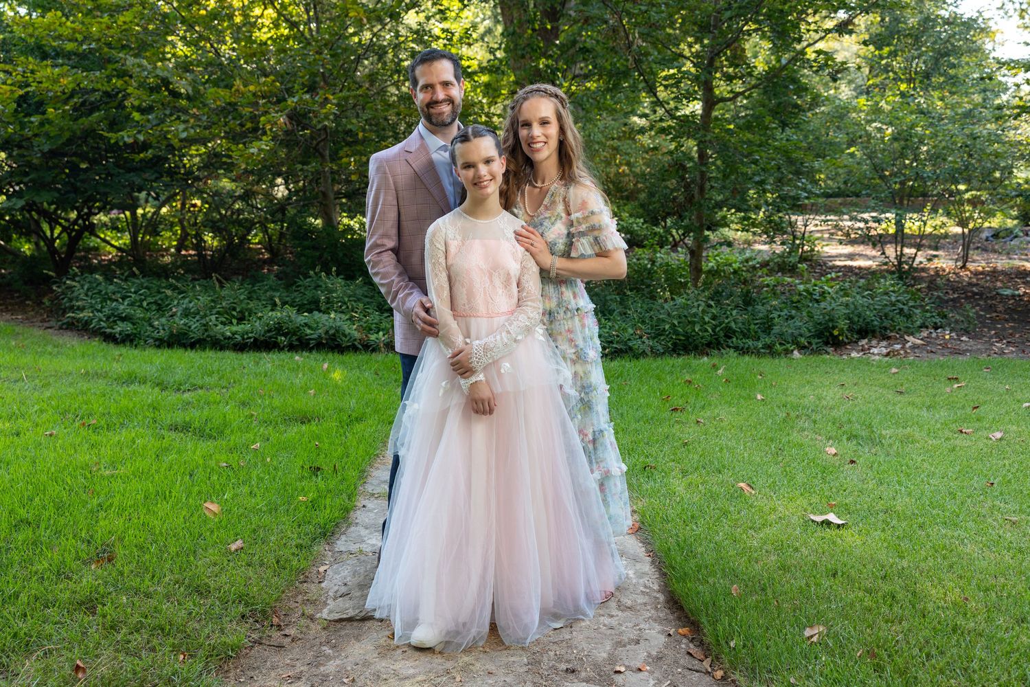 Three elegantly dressed people pose outdoors in formal attire, including pink and floral gowns, on a garden path.