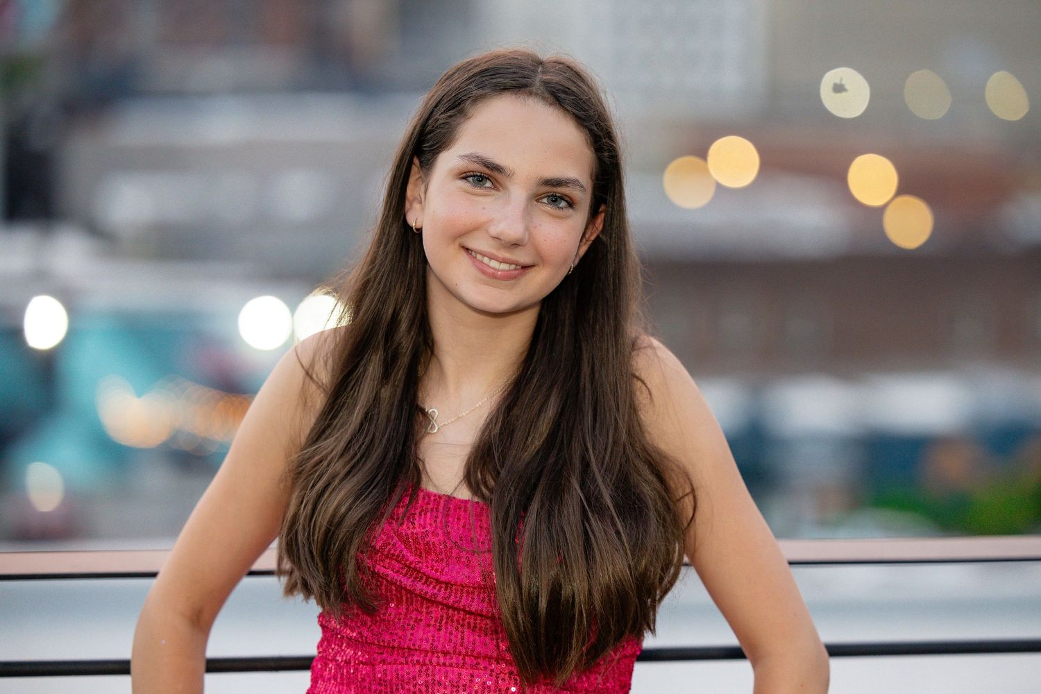 Young woman with long dark hair smiling in a pink sequin top, posing outdoors with bokeh city lights behind her.