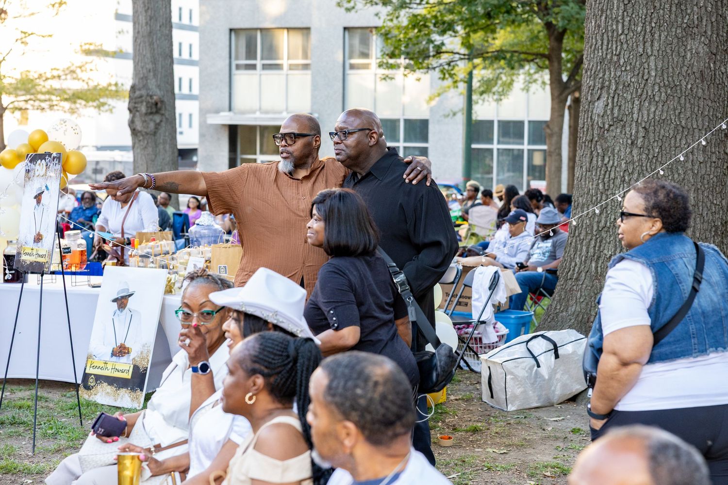 Outdoor community event with attendees sitting under trees on a sunny day.