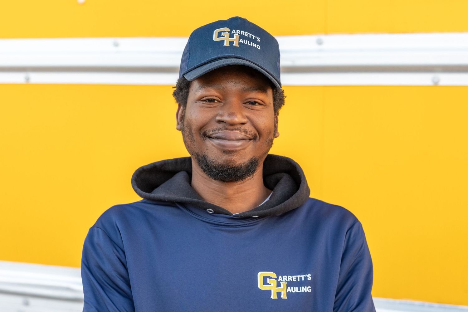 Team member wearing dark hoodie and cap with company logo on yellow background.
