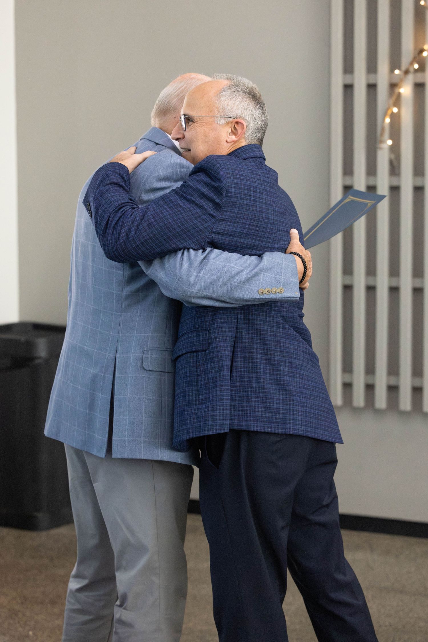 Two men in business suits embrace warmly in what appears to be an office hallway setting.