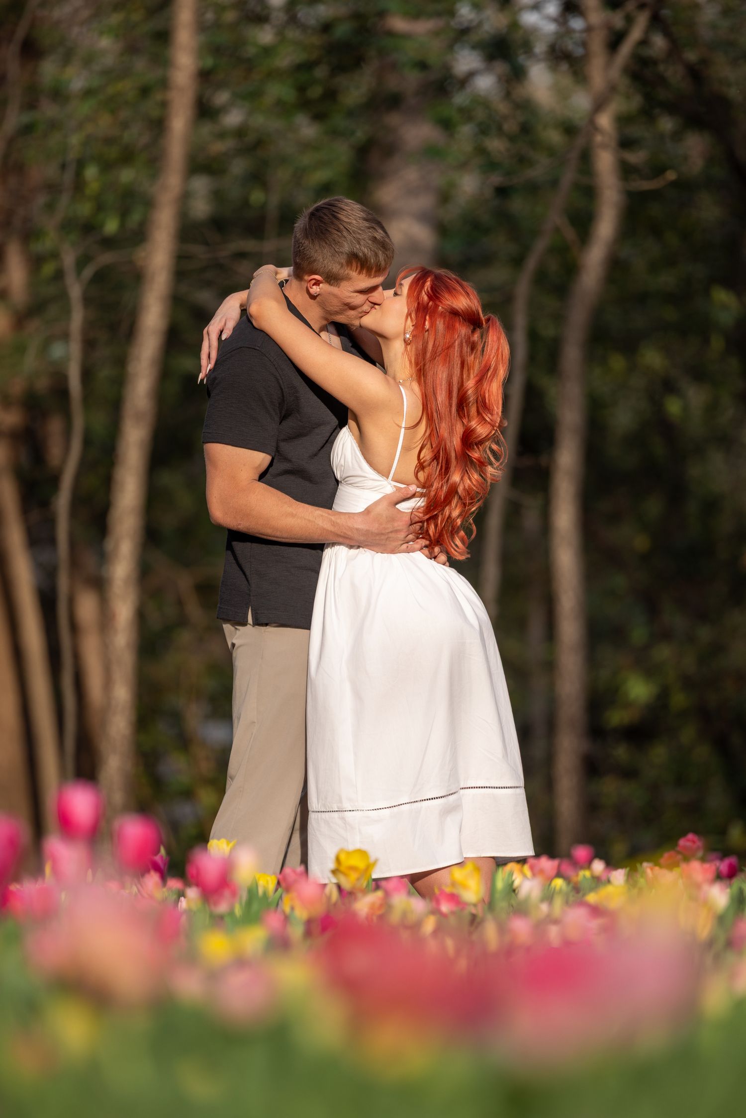 A romantic couple embraces in a garden with colorful flowers blooming in the foreground and trees in the background.
