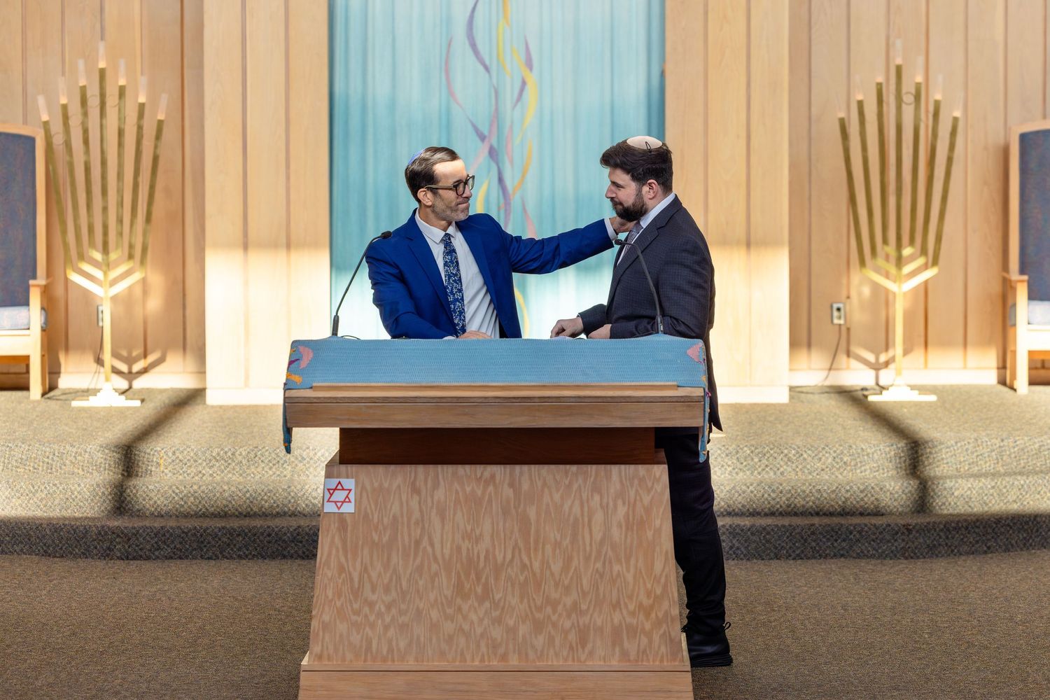 Two men shaking hands at a podium in a synagogue with menorahs and decorative Torah ark in background.