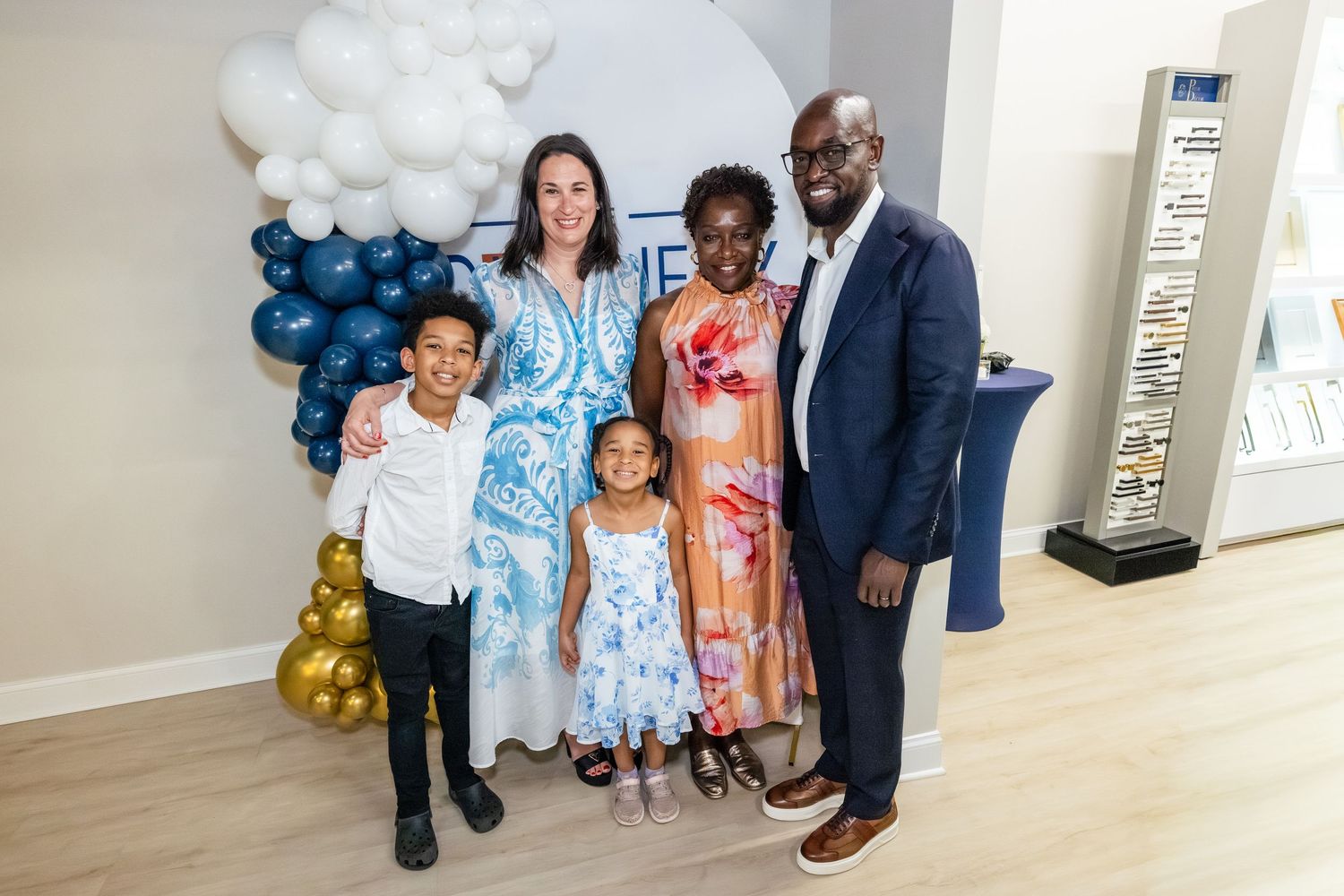 Family of five posing together at a celebratory event with balloon decorations in blue, white, and gold.