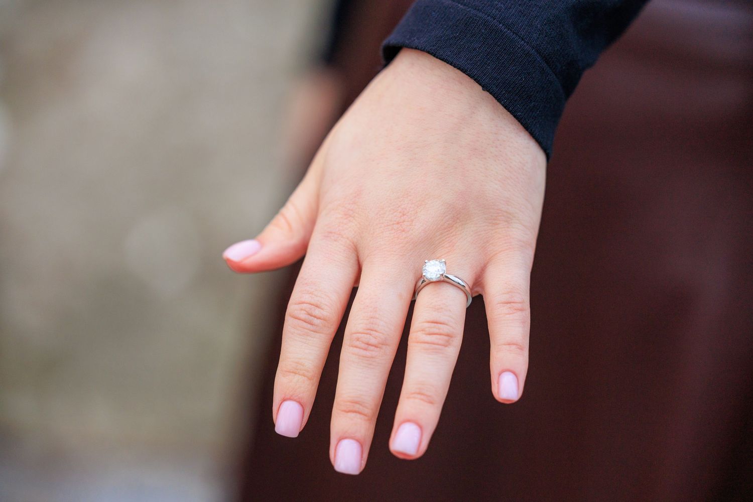 A hand wearing a sparkling engagement ring with pink painted fingernails against a blurred background.