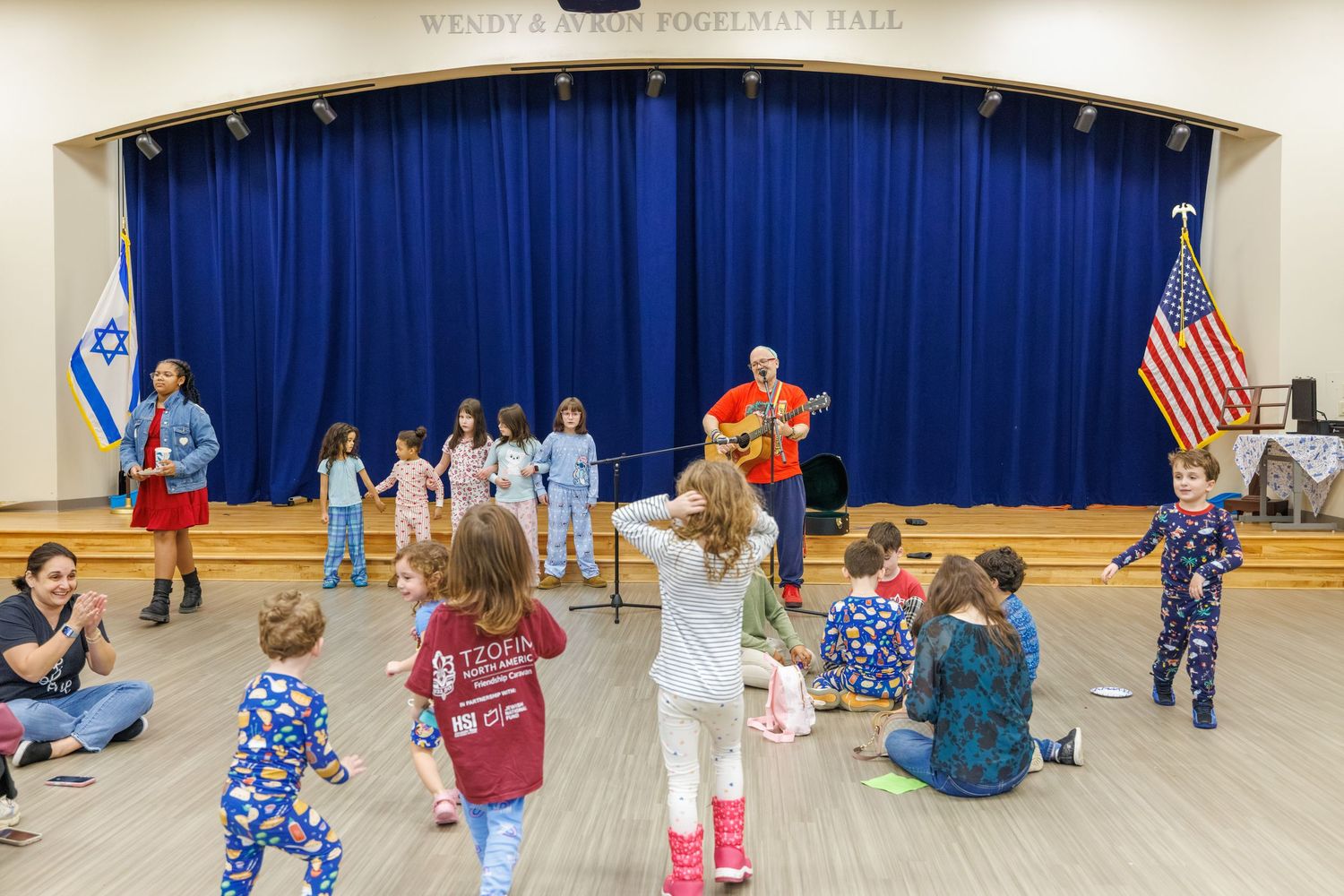 Children engage in group activities on a gym floor with American flag and blue stage curtains visible.