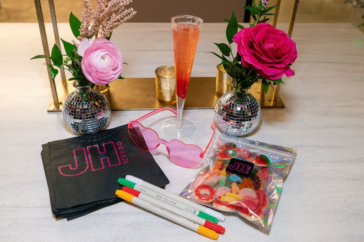 Bachelorette party setup with monogrammed napkins, pink sunglasses, champagne, disco balls, and floral arrangements.