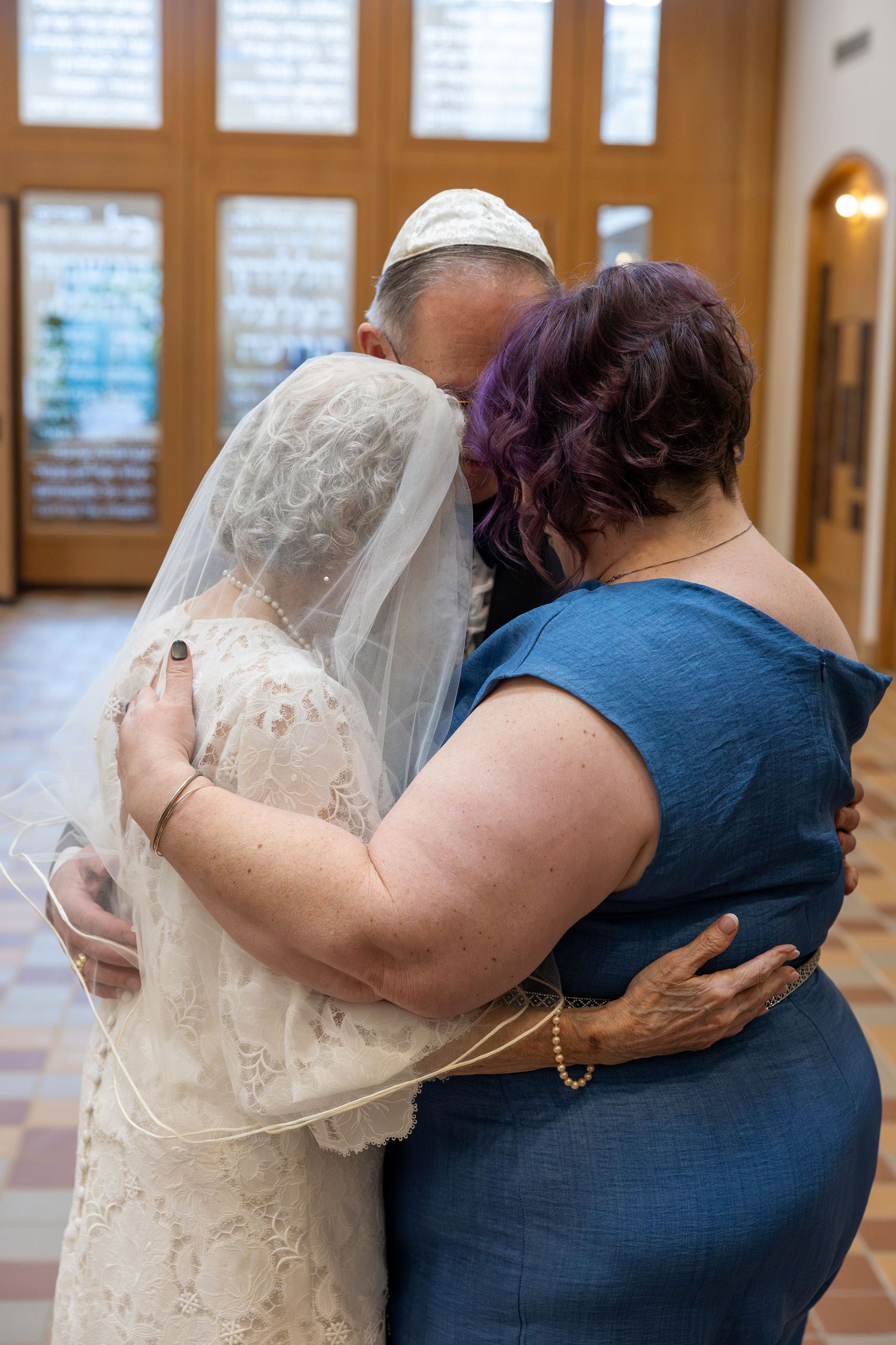 Two people embrace in a tender moment at a wedding, with one wearing a white lace dress and veil.