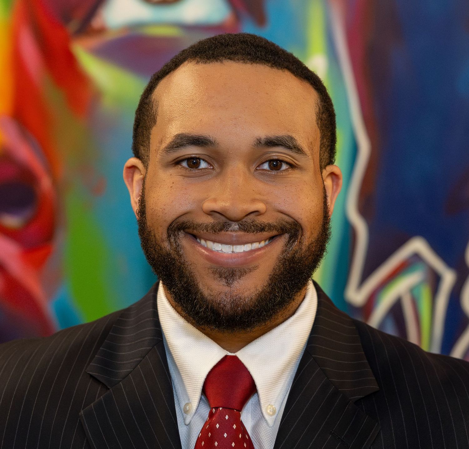 Professional headshot of a smiling person wearing a dark suit and red tie against a colorful abstract background.