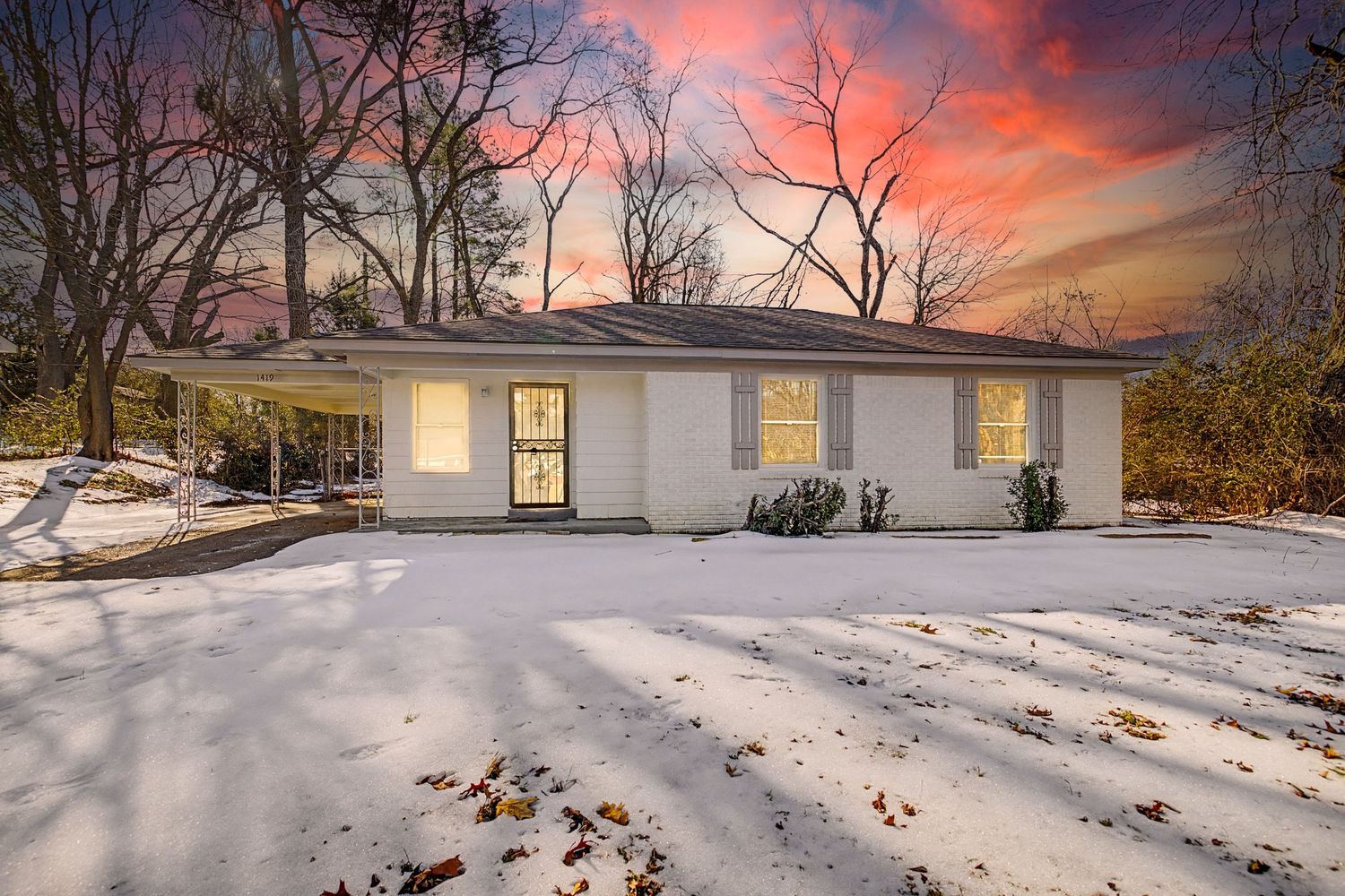 Snow-covered ranch home glows warmly at sunset with vibrant pink and orange sky, surrounded by bare winter trees.