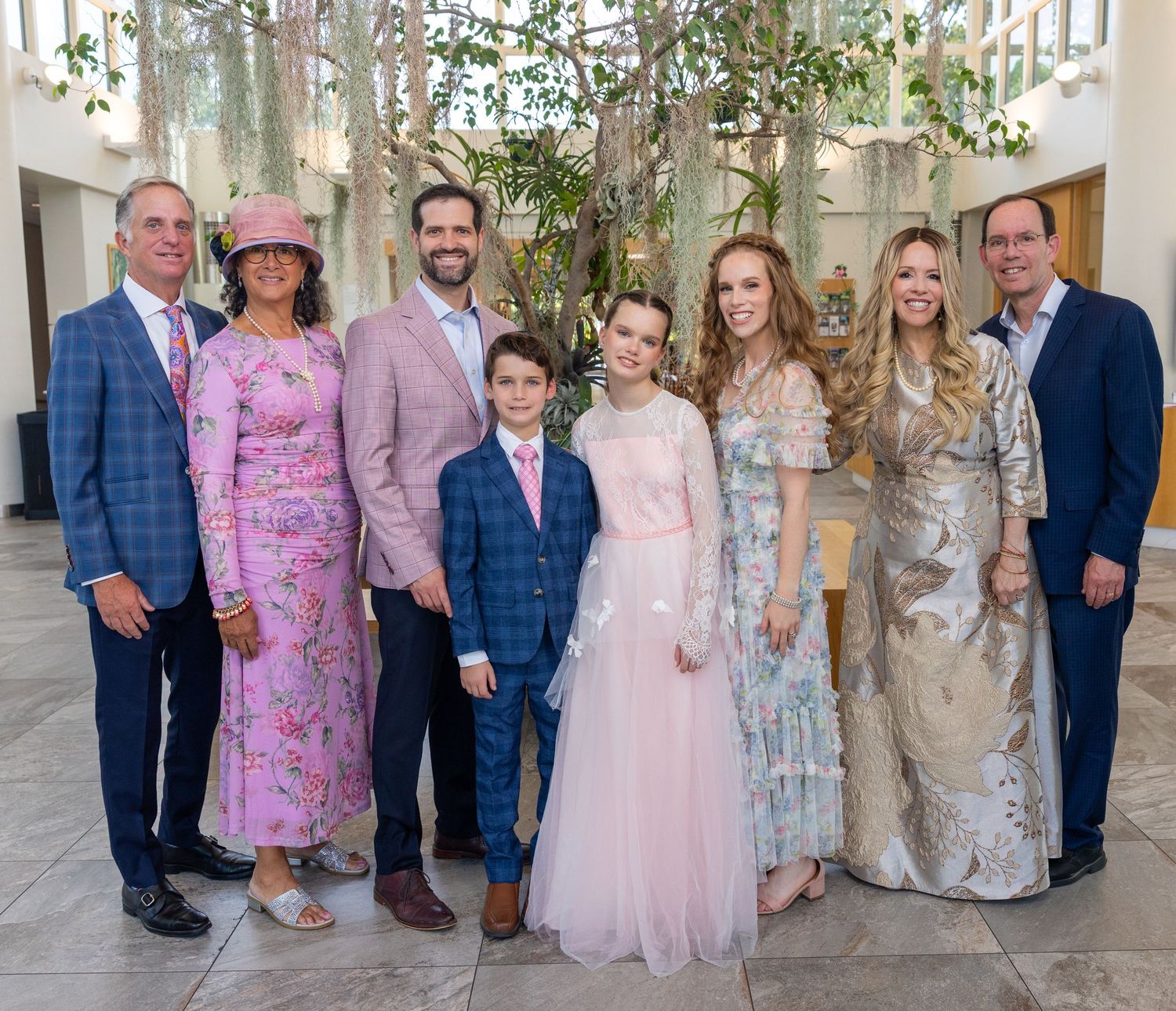 A joyful family group of eight poses together in formal attire at an elegant indoor venue with lush greenery.
