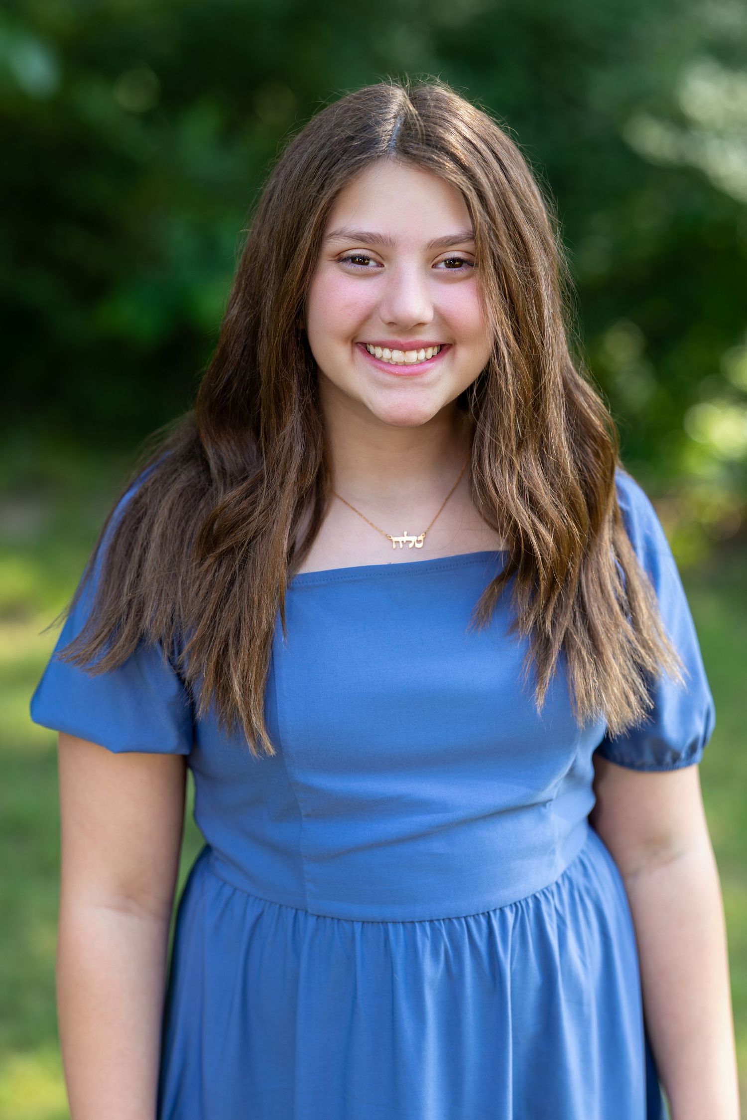 Smiling teenage girl in blue dress with necklace posing outdoors with green foliage background.