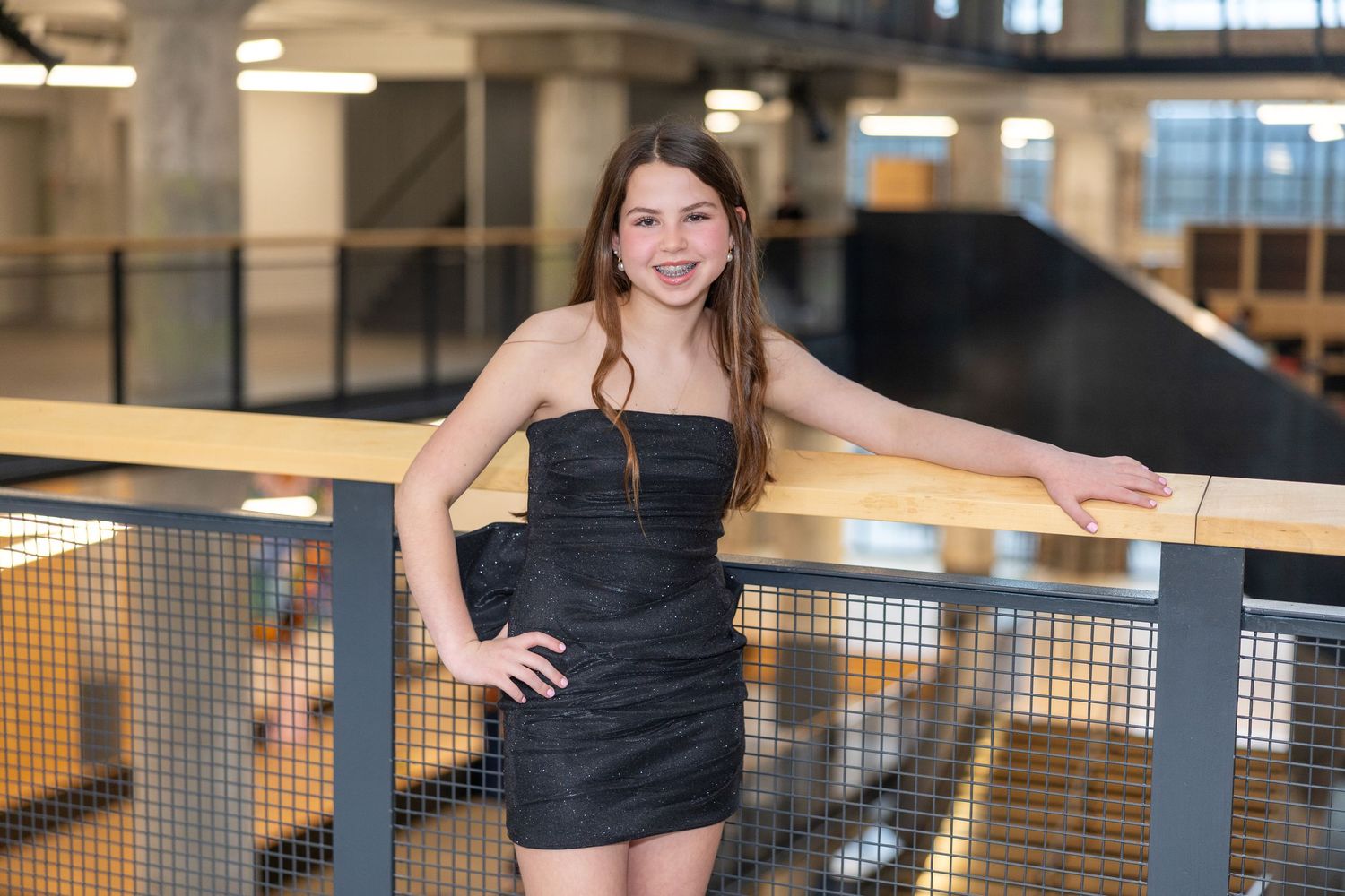 Young woman in black strapless mini dress posing confidently against railing in modern industrial building.
