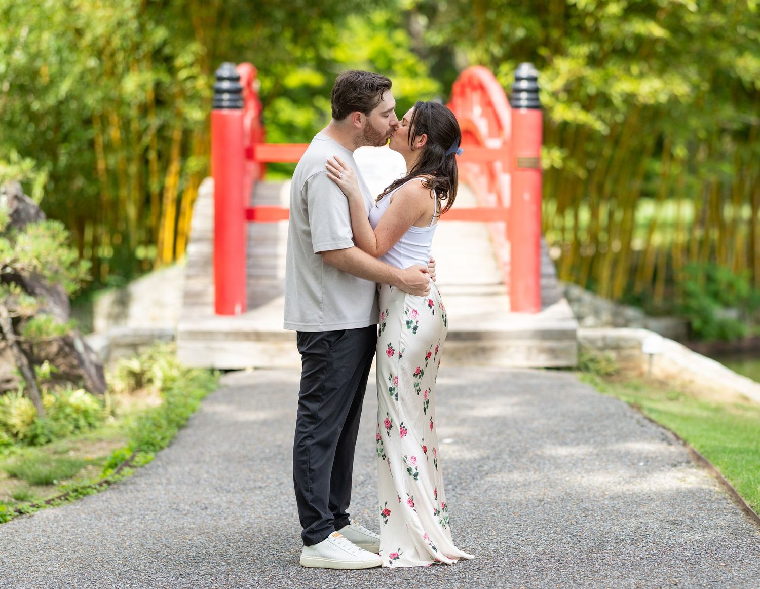 Couple sharing a romantic kiss on a garden path with a red bridge and bamboo background.
