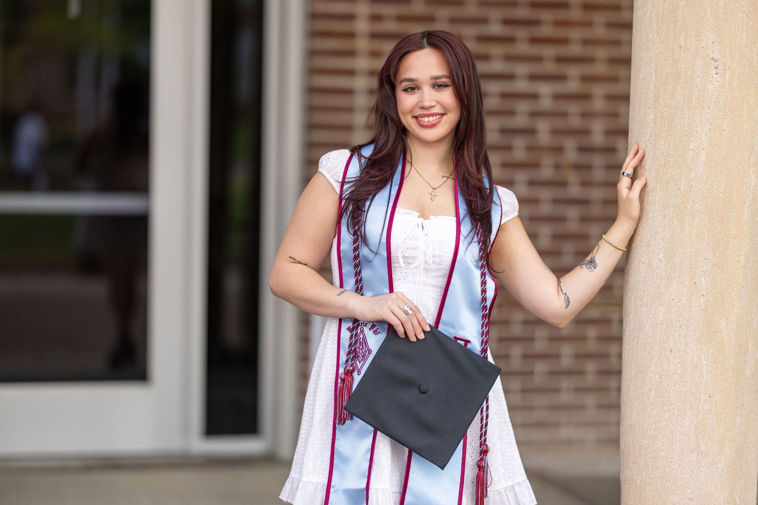 Smiling female graduate in white dress holding cap with colorful sash poses outdoors near building columns.
