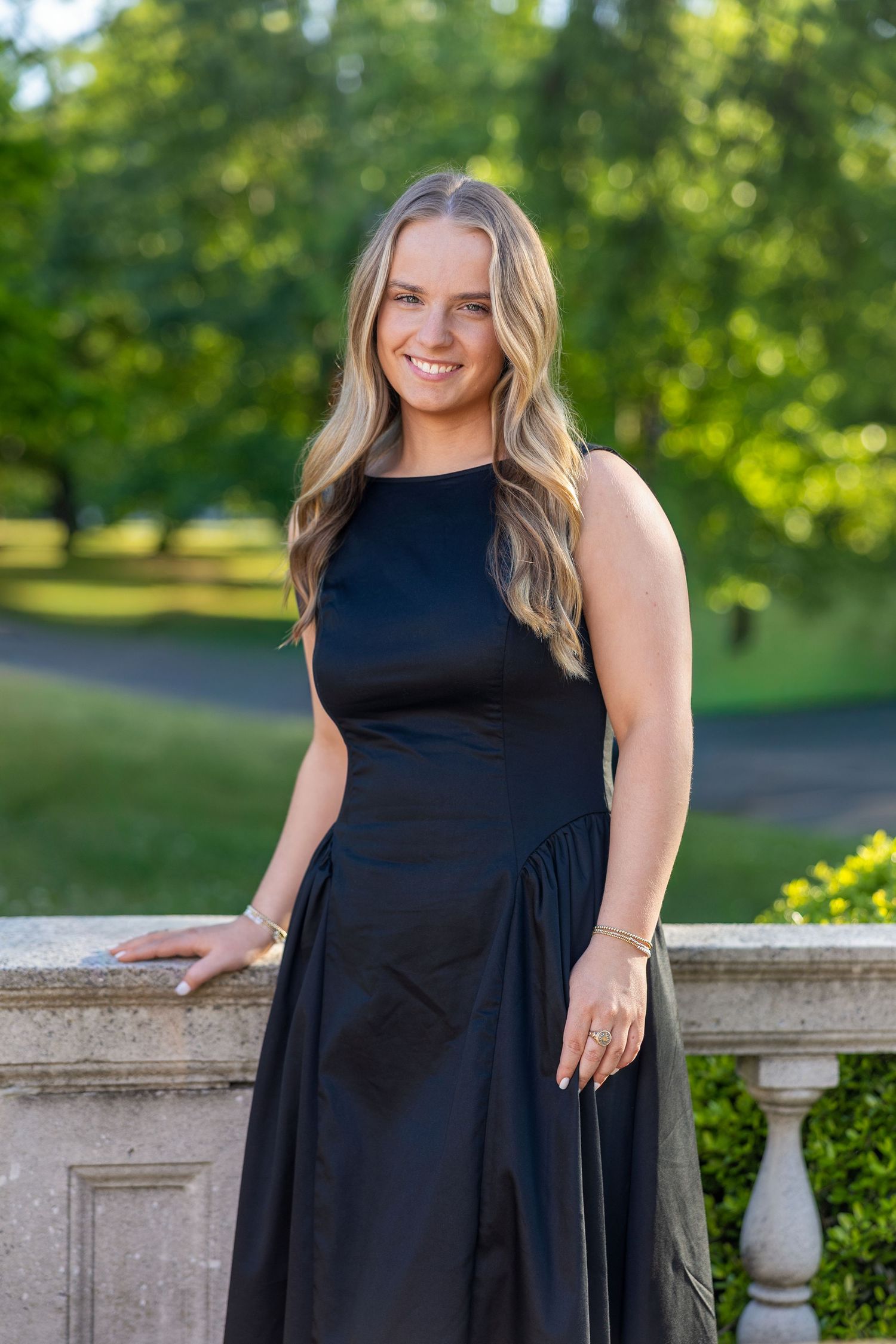 Young woman in black sleeveless maxi dress smiling outdoors by stone balustrade with lush green trees.