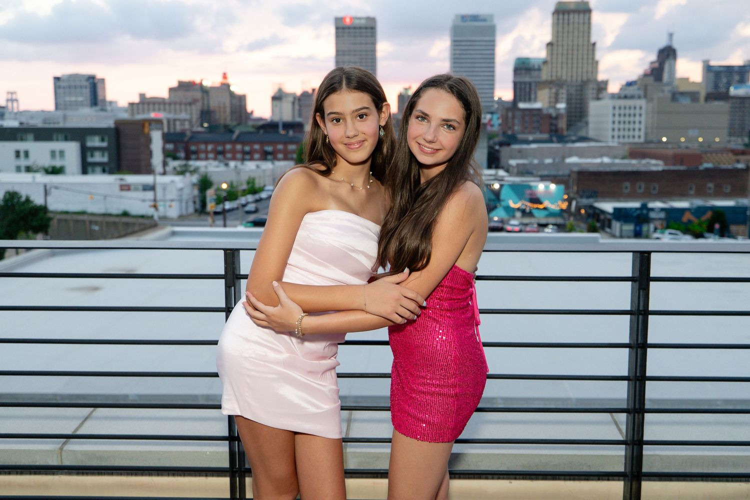 Two young women in elegant mini dresses pose together on a rooftop with a city skyline at dusk.