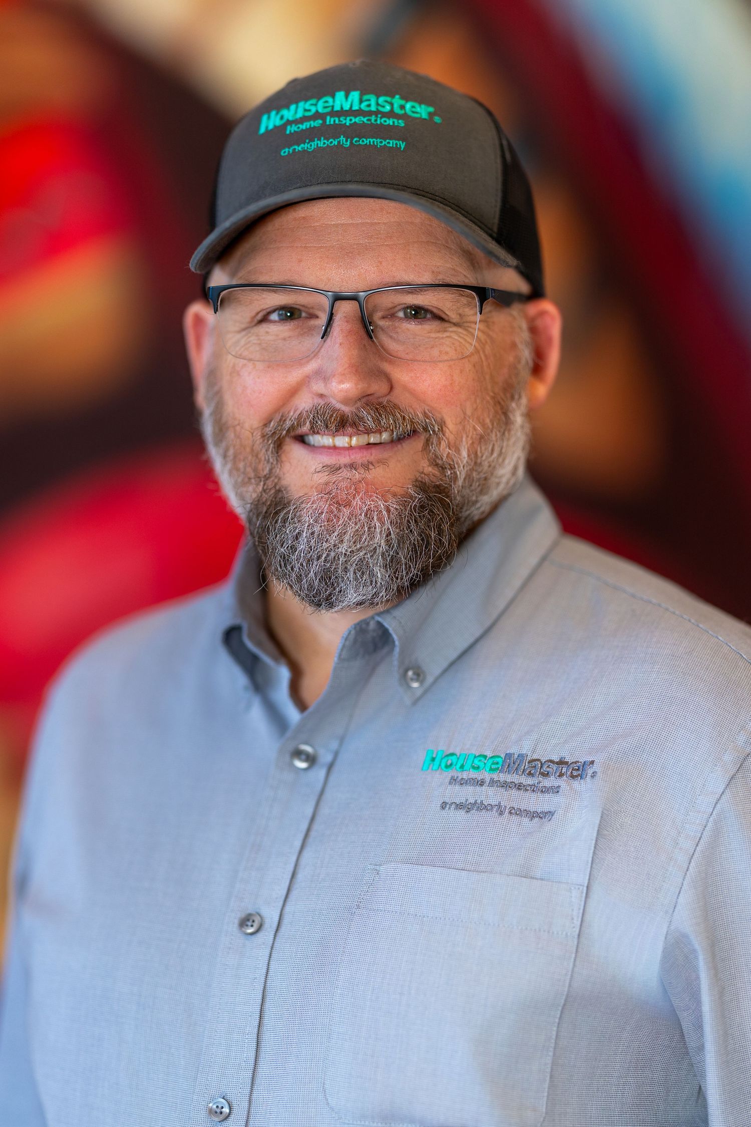 HouseMaster home inspector smiling in branded cap and shirt, wearing glasses with a neat beard.