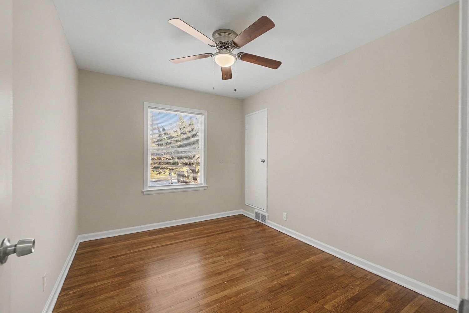Empty bedroom with dark hardwood floors, ceiling fan, and a window with natural light in a neutral-toned room.