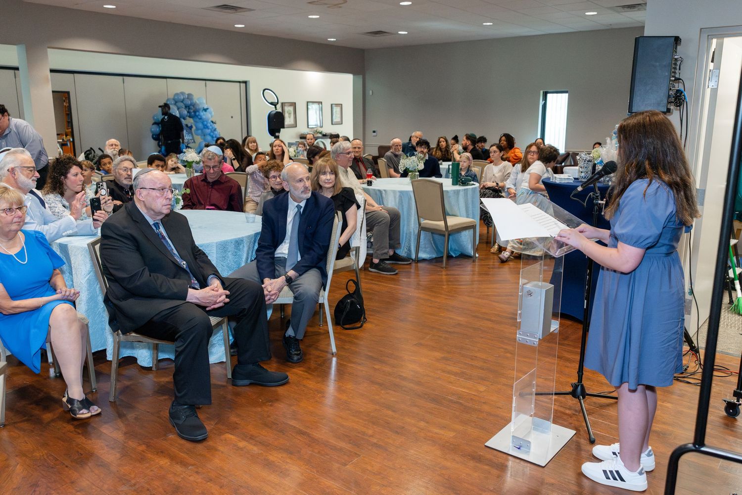 Woman in blue dress speaks at podium during formal indoor event with seated audience in decorated banquet hall.