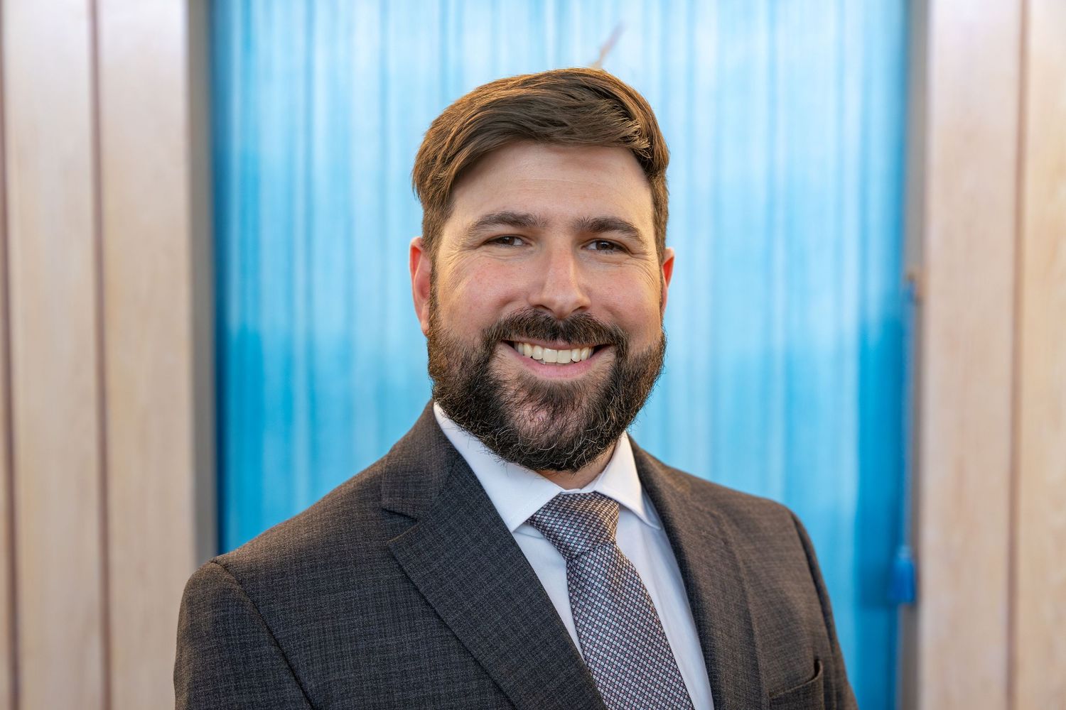 Professional headshot of a smiling bearded man in a gray suit and tie against a blue background.