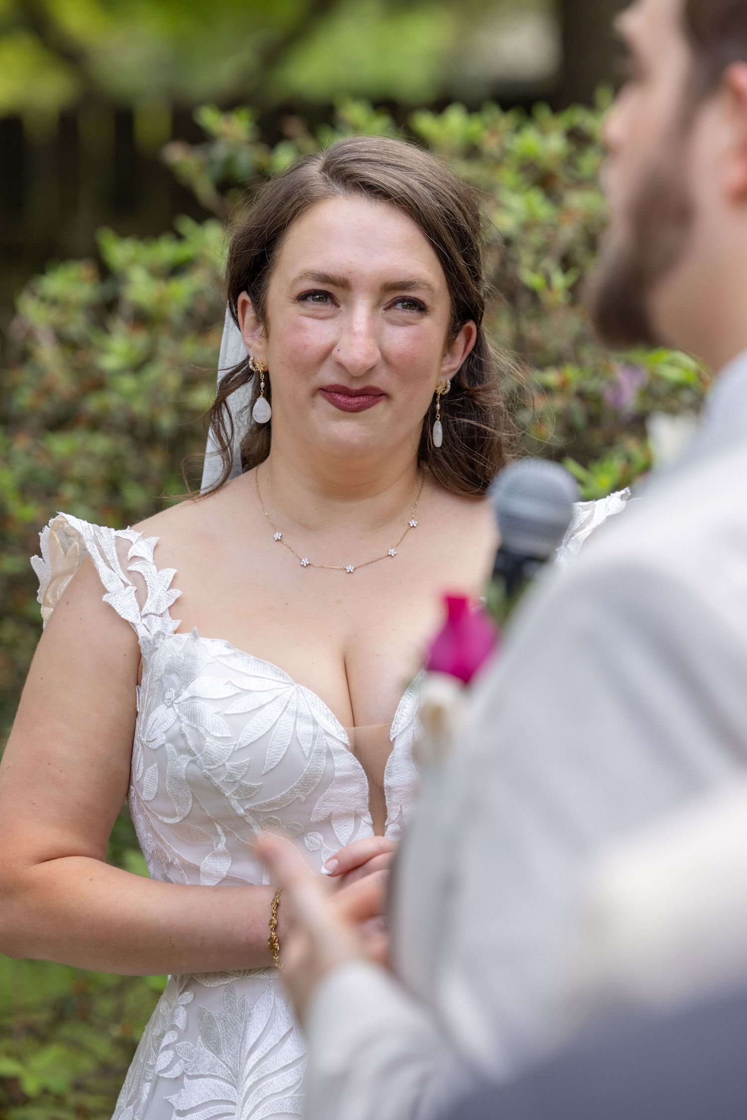 Emotional bride in white lace gown with veil exchanges vows during outdoor wedding ceremony.