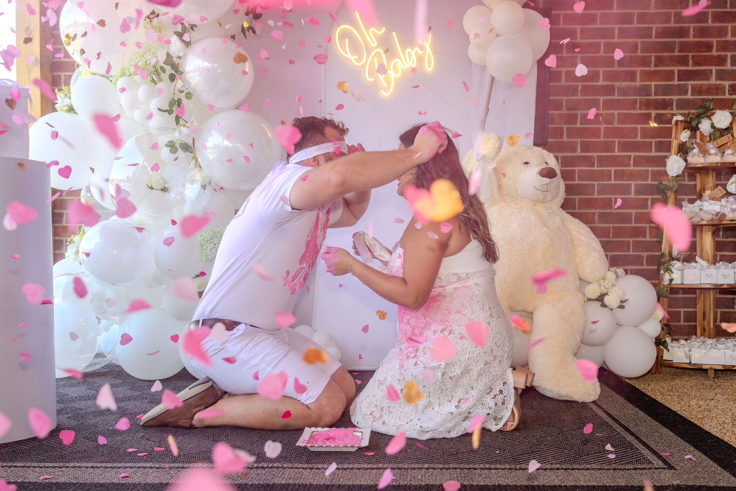Pink confetti and white balloons create a festive atmosphere against a brick wall backdrop.