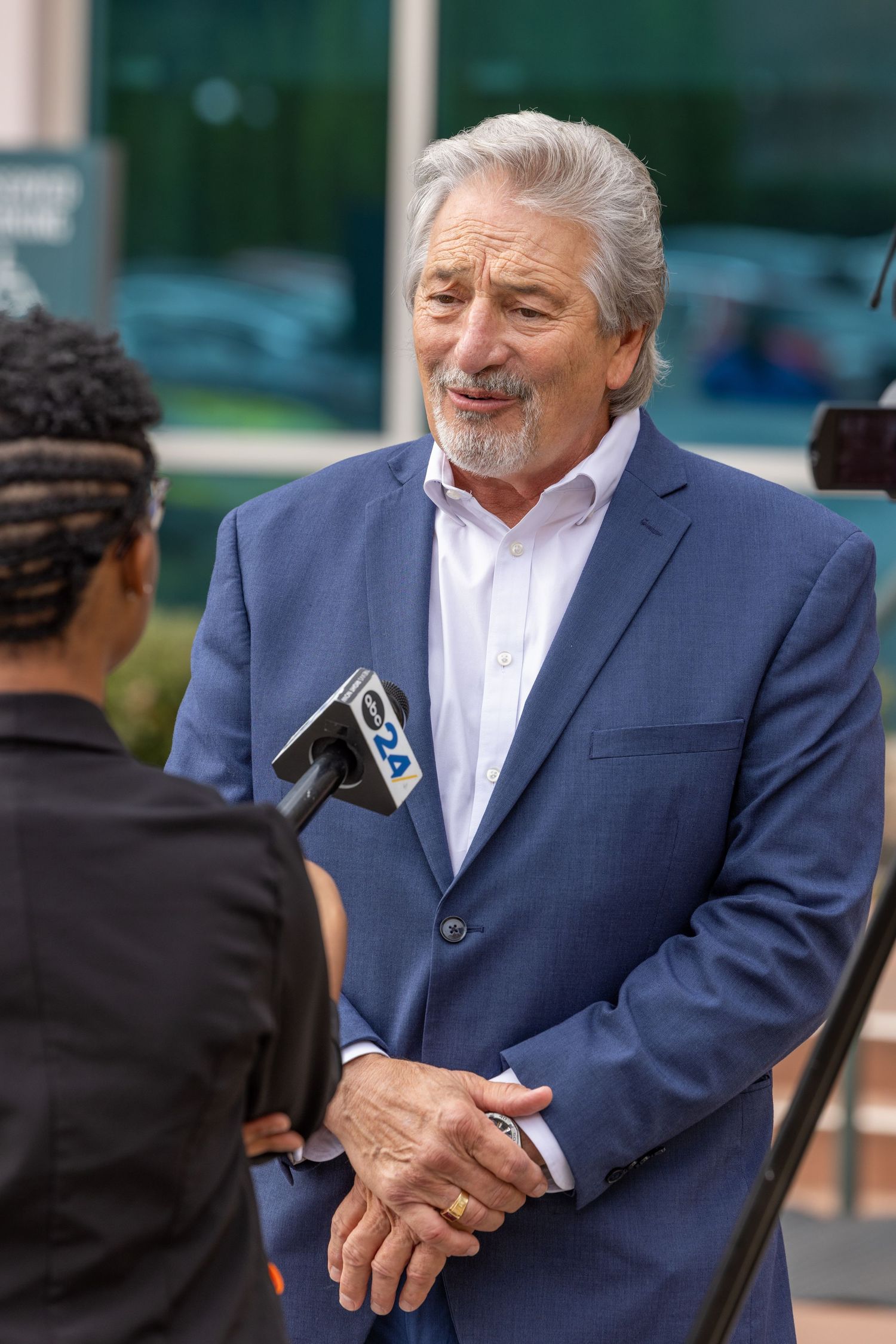 Man in navy suit speaking at outdoor corporate gathering.