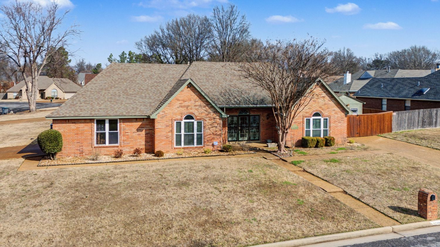 Aerial view of a single-story brick ranch home with tan roof and dormant winter lawn in suburban neighborhood.