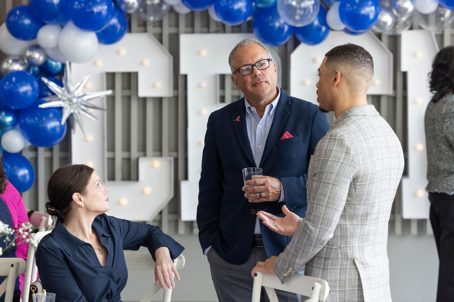 Professional colleagues engage in conversation at a business event decorated with blue balloons and modern decor.