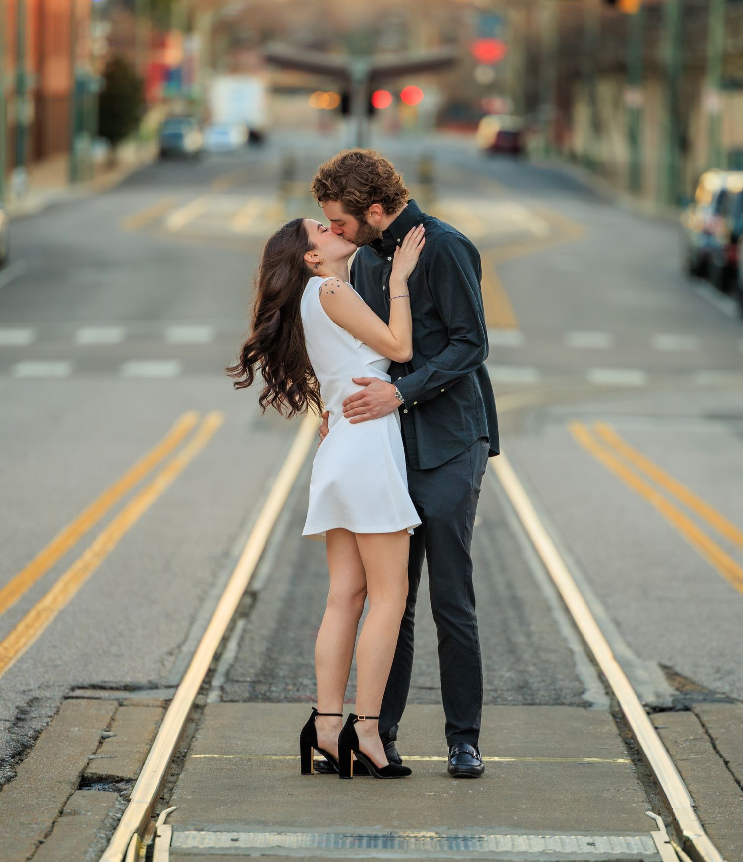 A romantic couple shares an intimate moment while standing on train tracks in an urban setting.