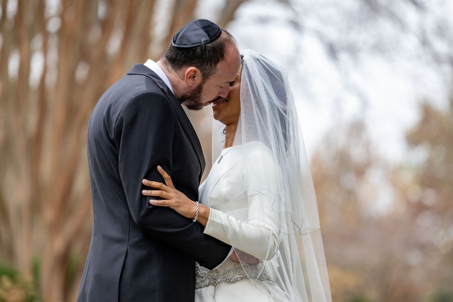 Romantic wedding couple embraces outdoors in autumn with bride wearing flowing white veil and groom in dark suit.