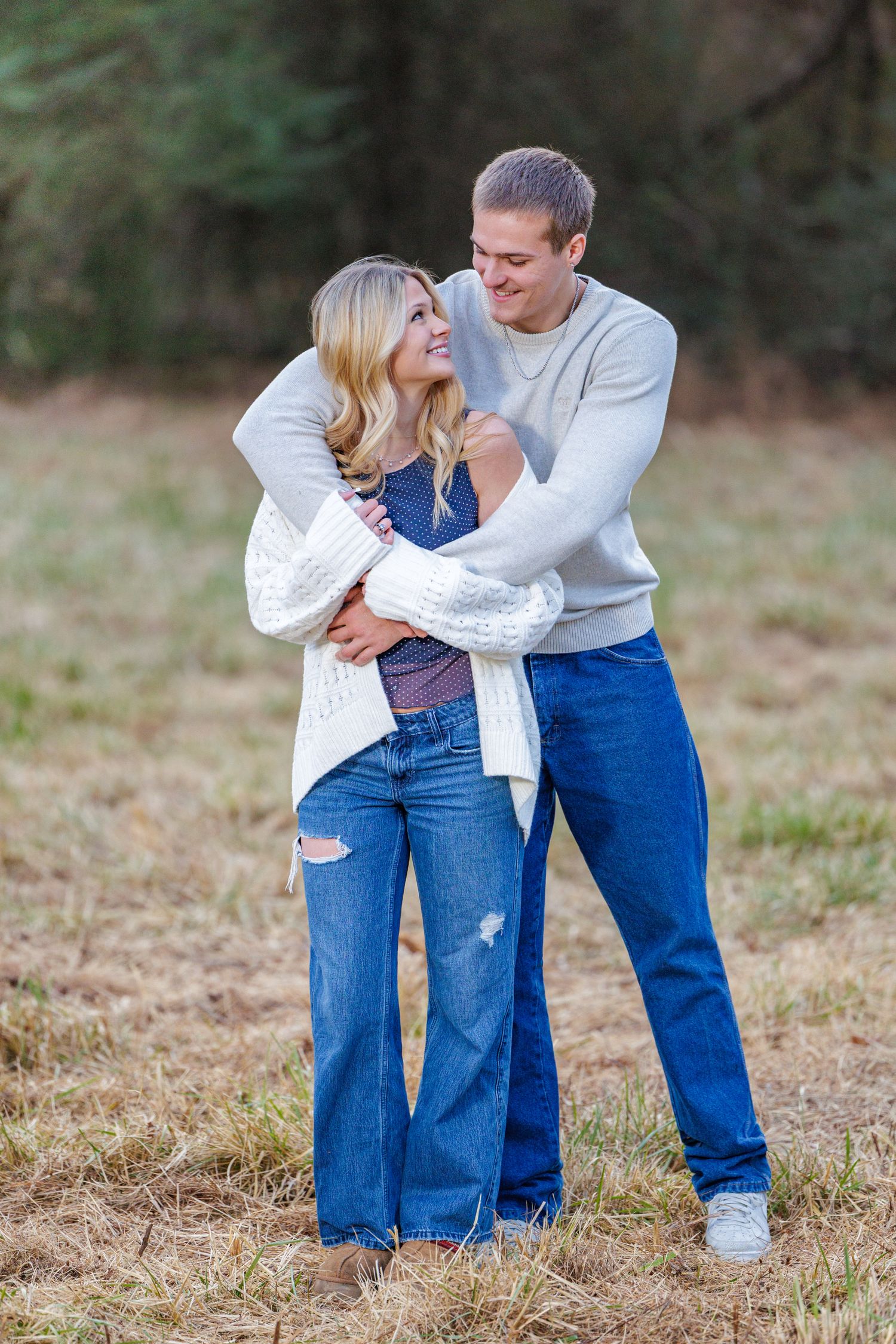 A couple in matching jeans and white tops share a tender embrace while standing in a grassy field.