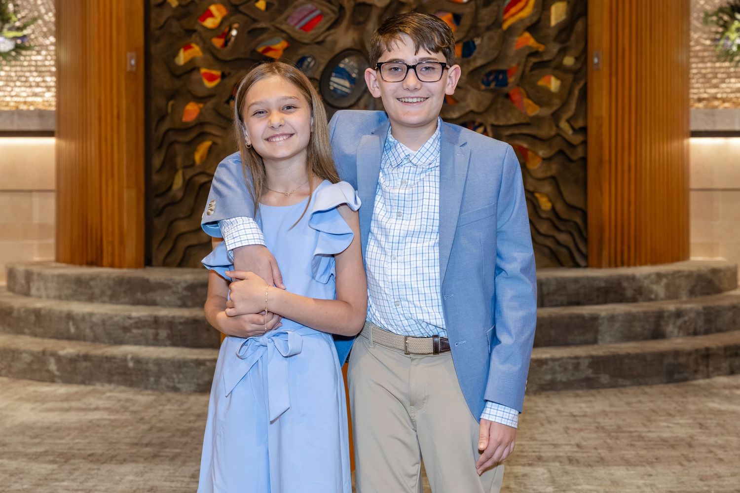 Two children dressed in formal blue outfits smiling together in an elegant lobby setting.