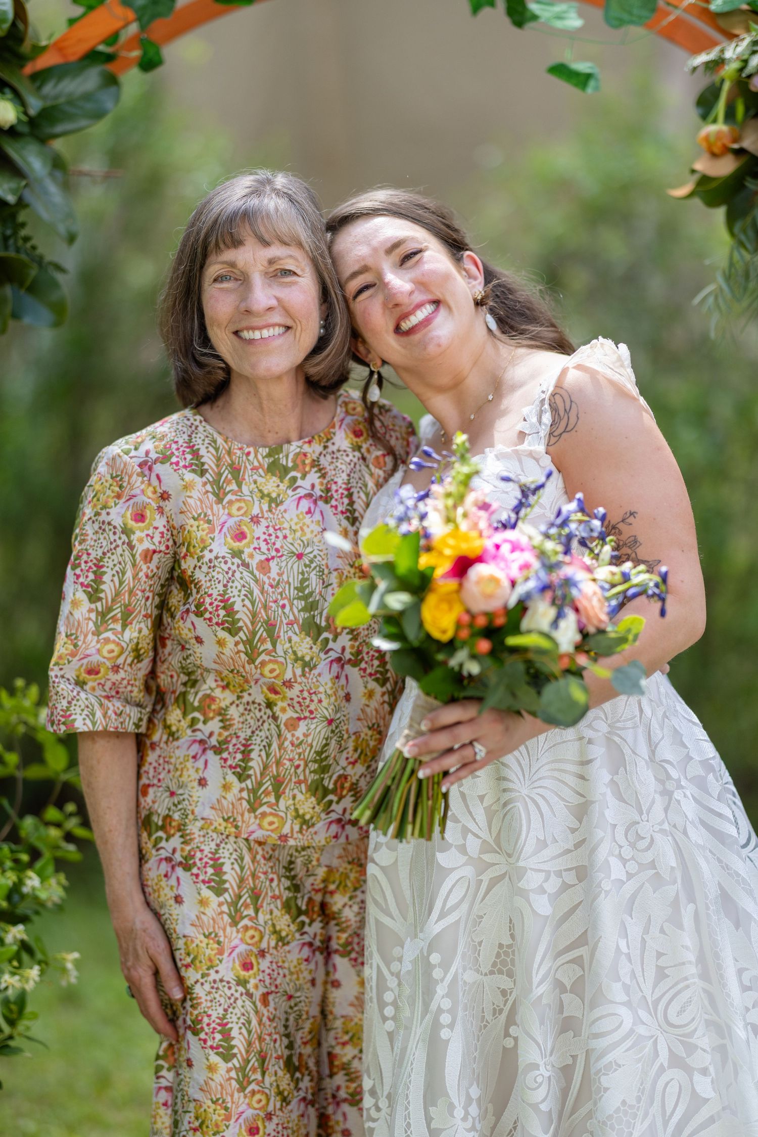 Bride in lace gown holds colorful bouquet beside smiling woman in floral dress at outdoor wedding ceremony.