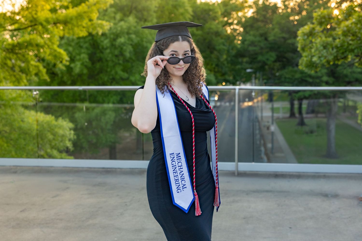 Young woman in graduation cap and gown with sash, posing outdoors while adjusting sunglasses on a sunny day.