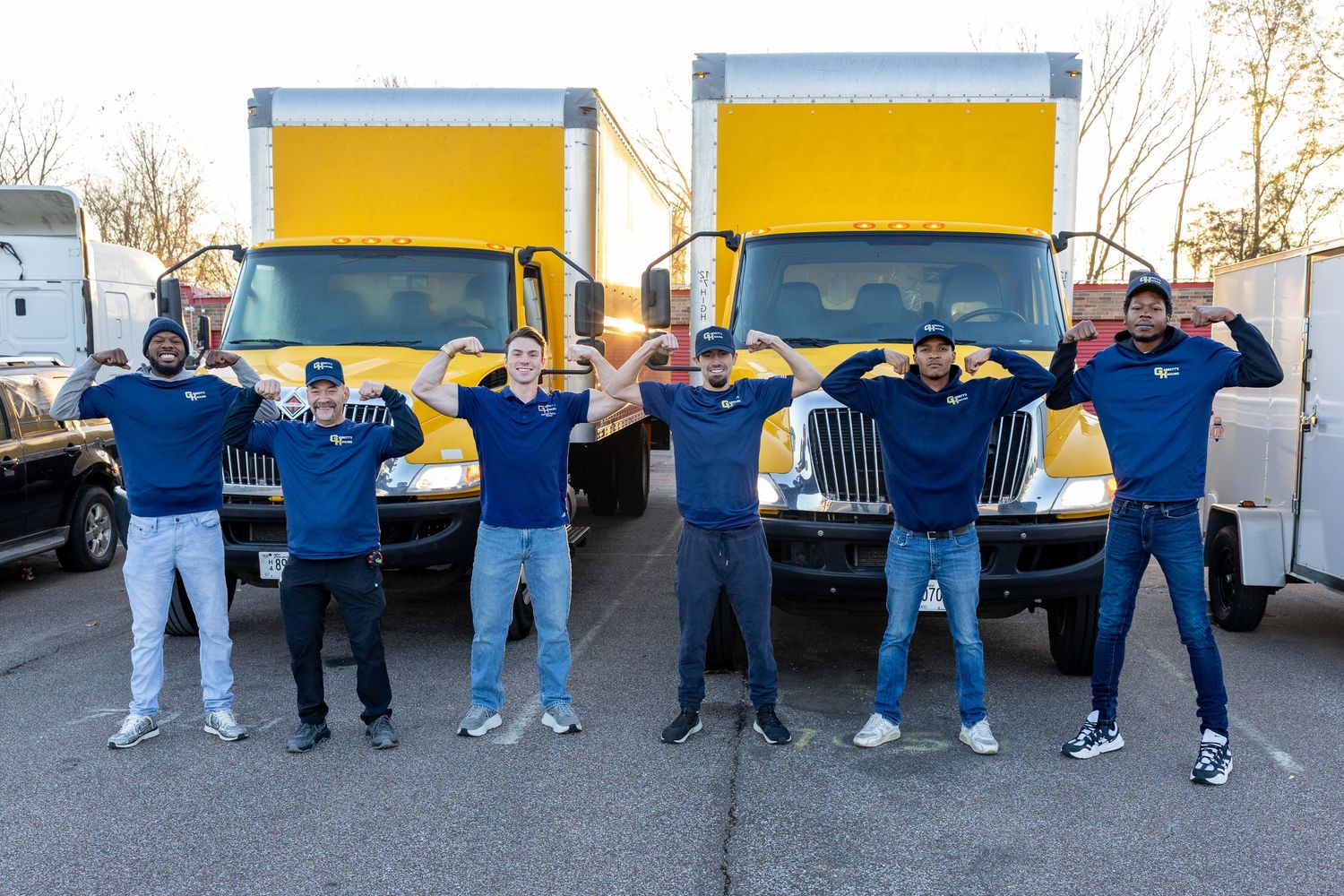 Group of delivery service workers lined up in front of yellow trucks in a parking lot.