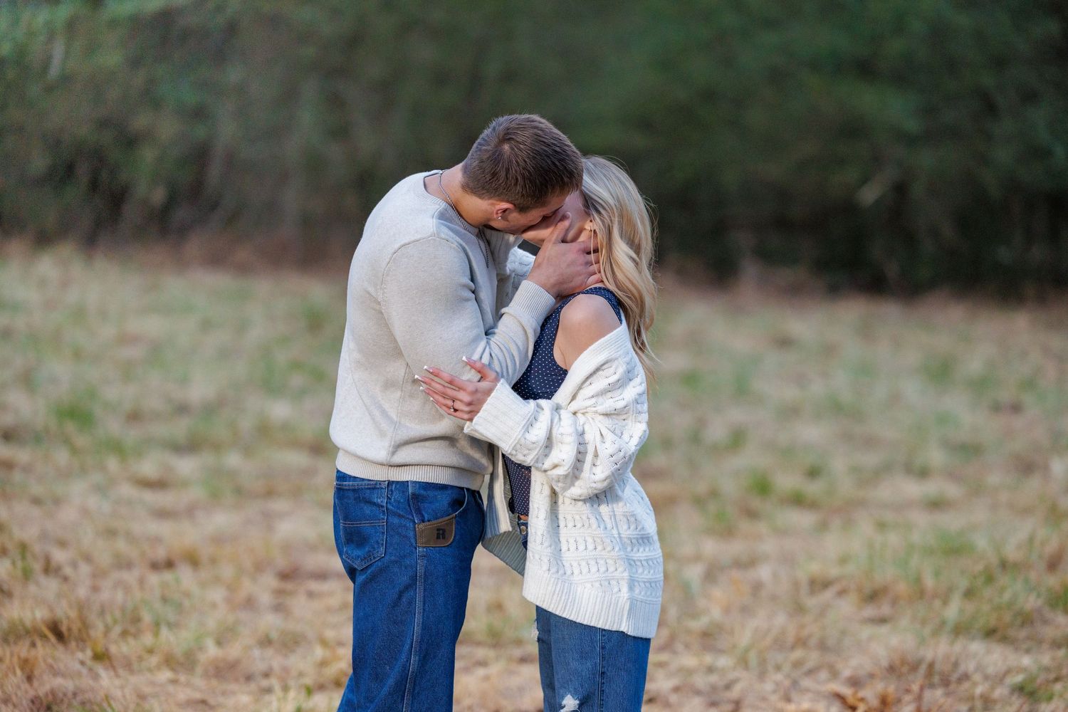 A romantic couple embraces in a field wearing cozy sweaters during an outdoor photo session.