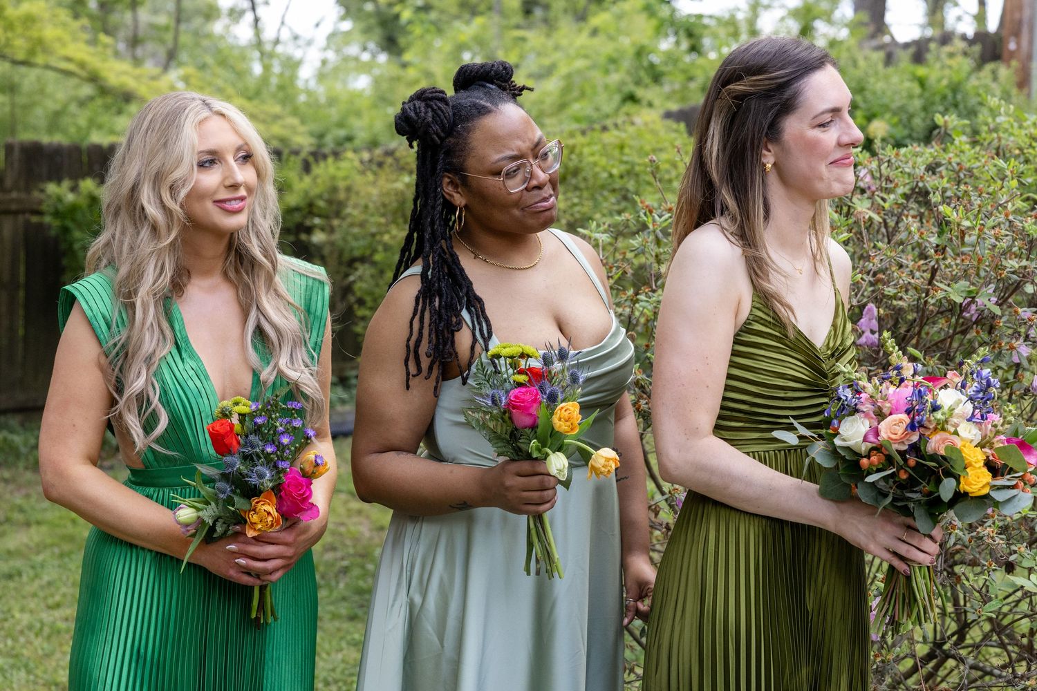 Three bridesmaids in green dresses holding colorful bouquets stand outdoors at a garden wedding ceremony.