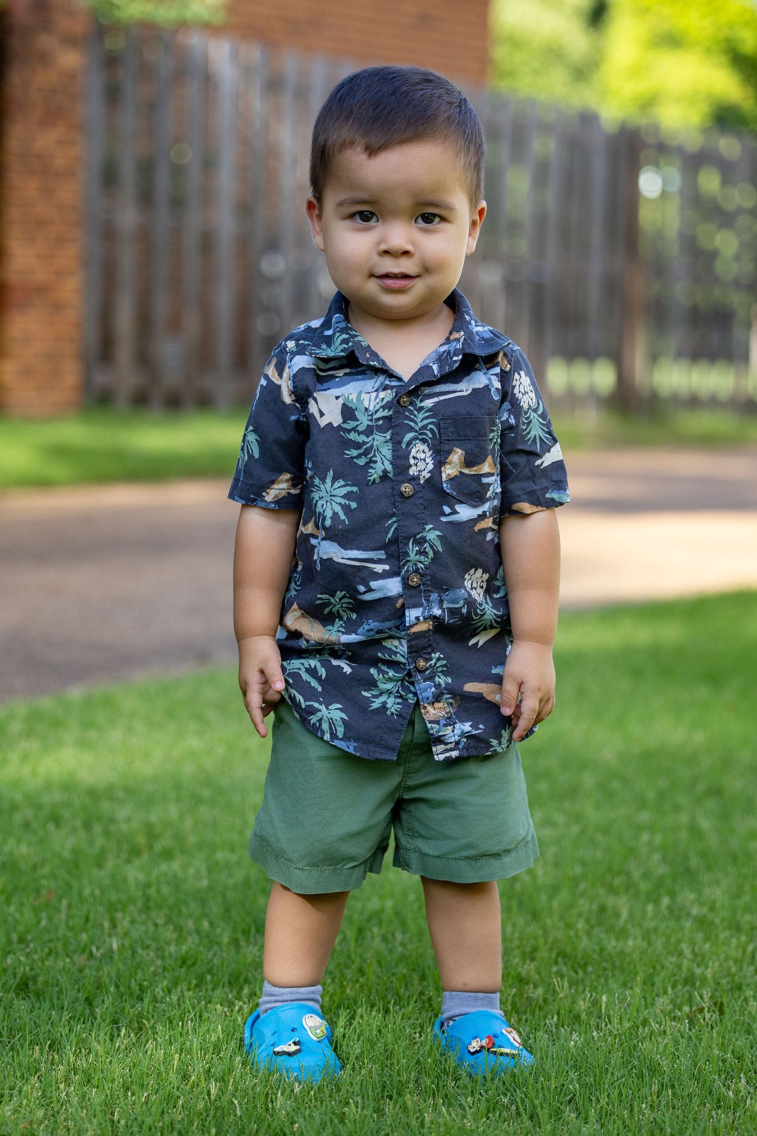 Toddler boy in tropical print shirt, green shorts, and blue Crocs standing on green grass in a backyard.