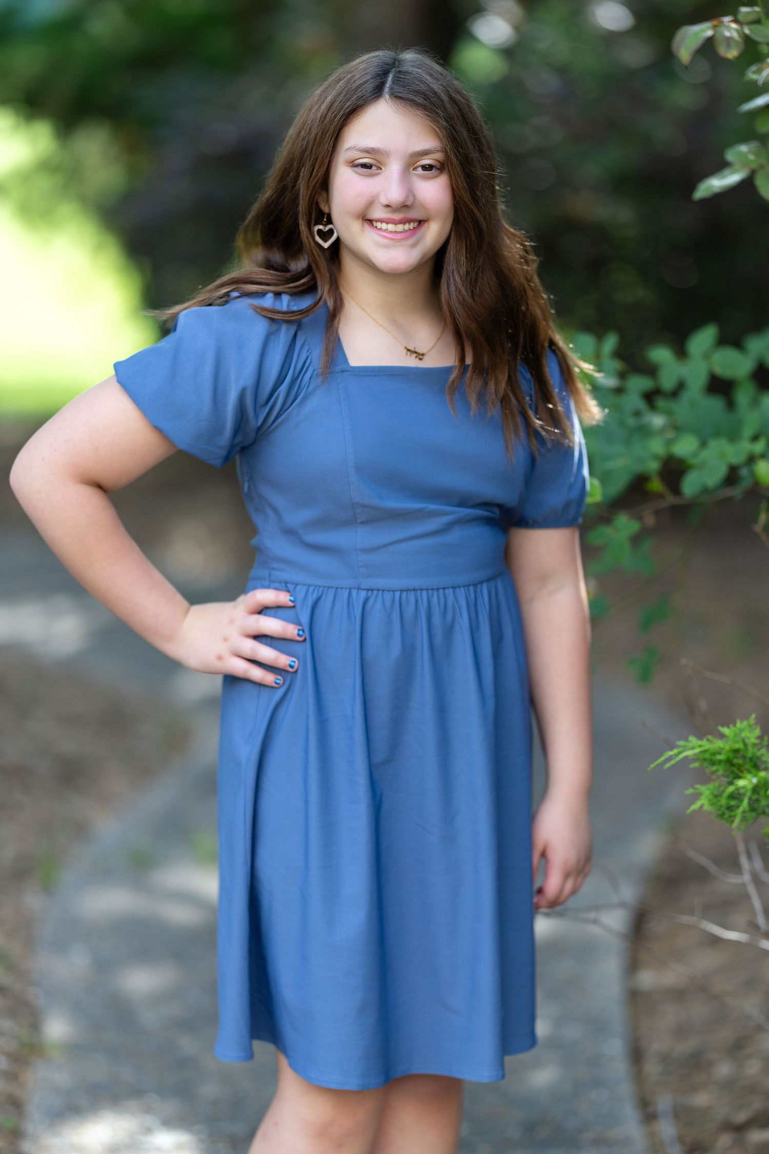 Smiling young girl in a blue square-neck dress posing outdoors on a garden path surrounded by greenery.