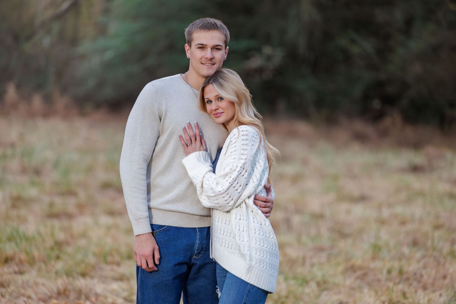 A couple in casual fall attire poses together for romantic outdoor engagement photos in a field at sunset.