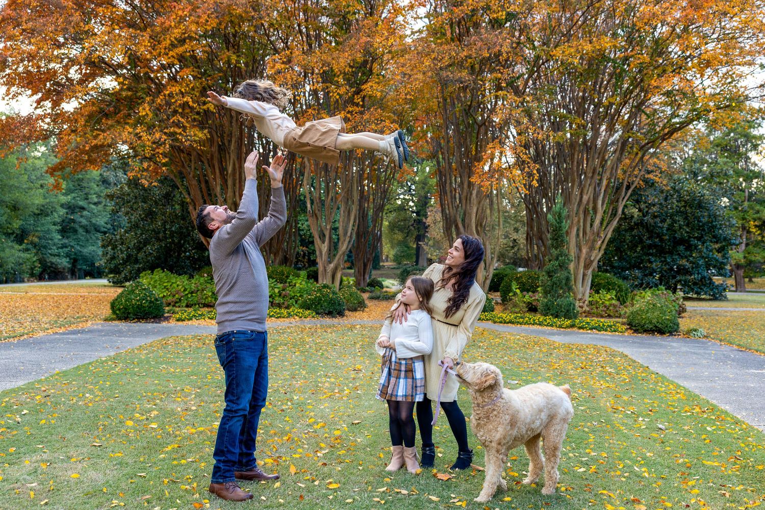 Family and dog enjoying autumn scenery with fallen leaves in a park setting.